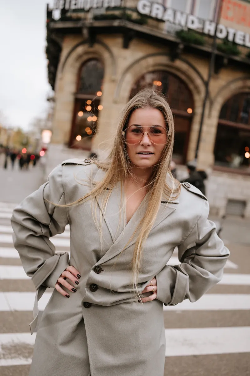 Clenda Moen met de handen in de zij in een beige trenchcoat voor het Grand Hotel