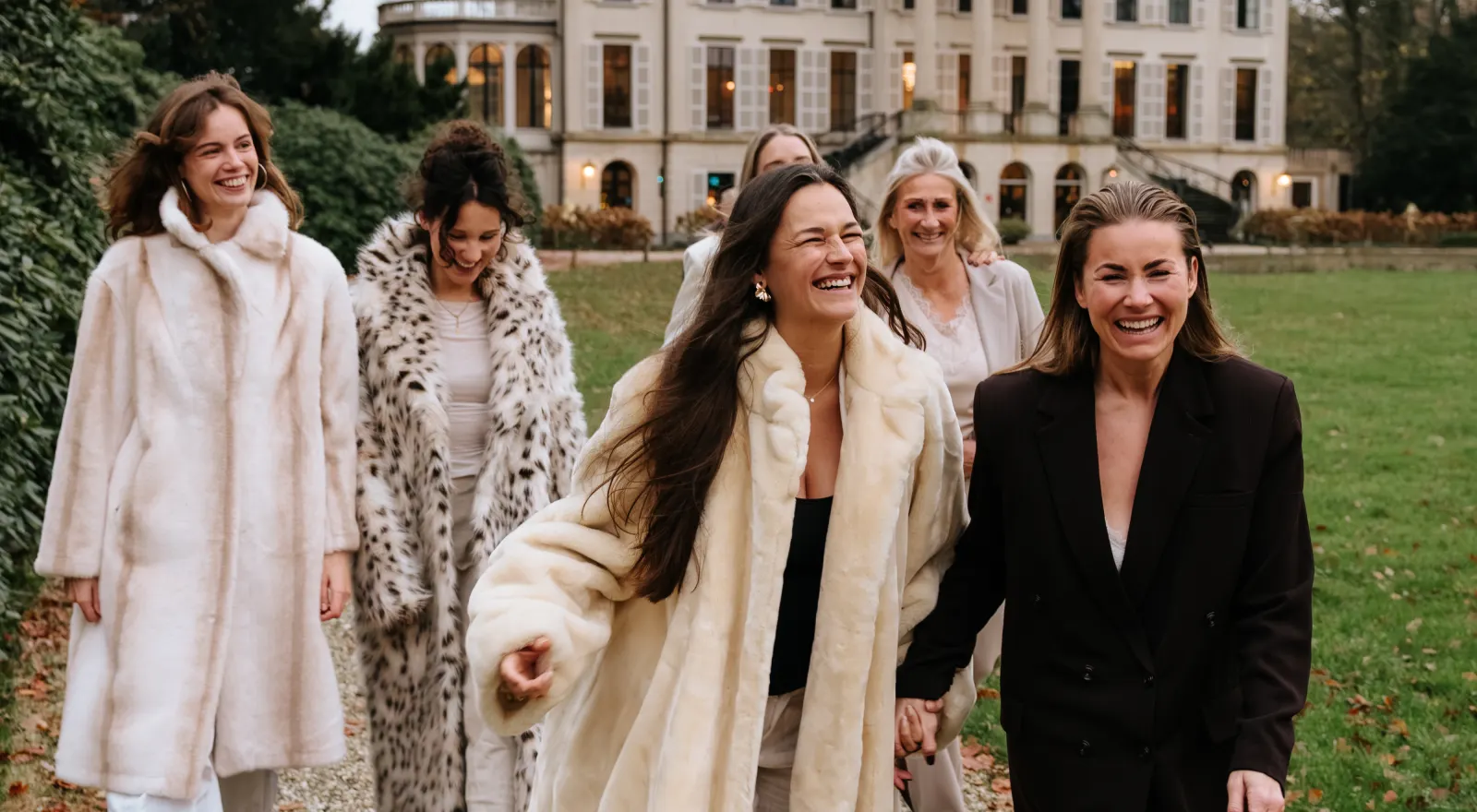 Een groep lachende vrouwen in lange jassen op een grasveld voor een landhuis